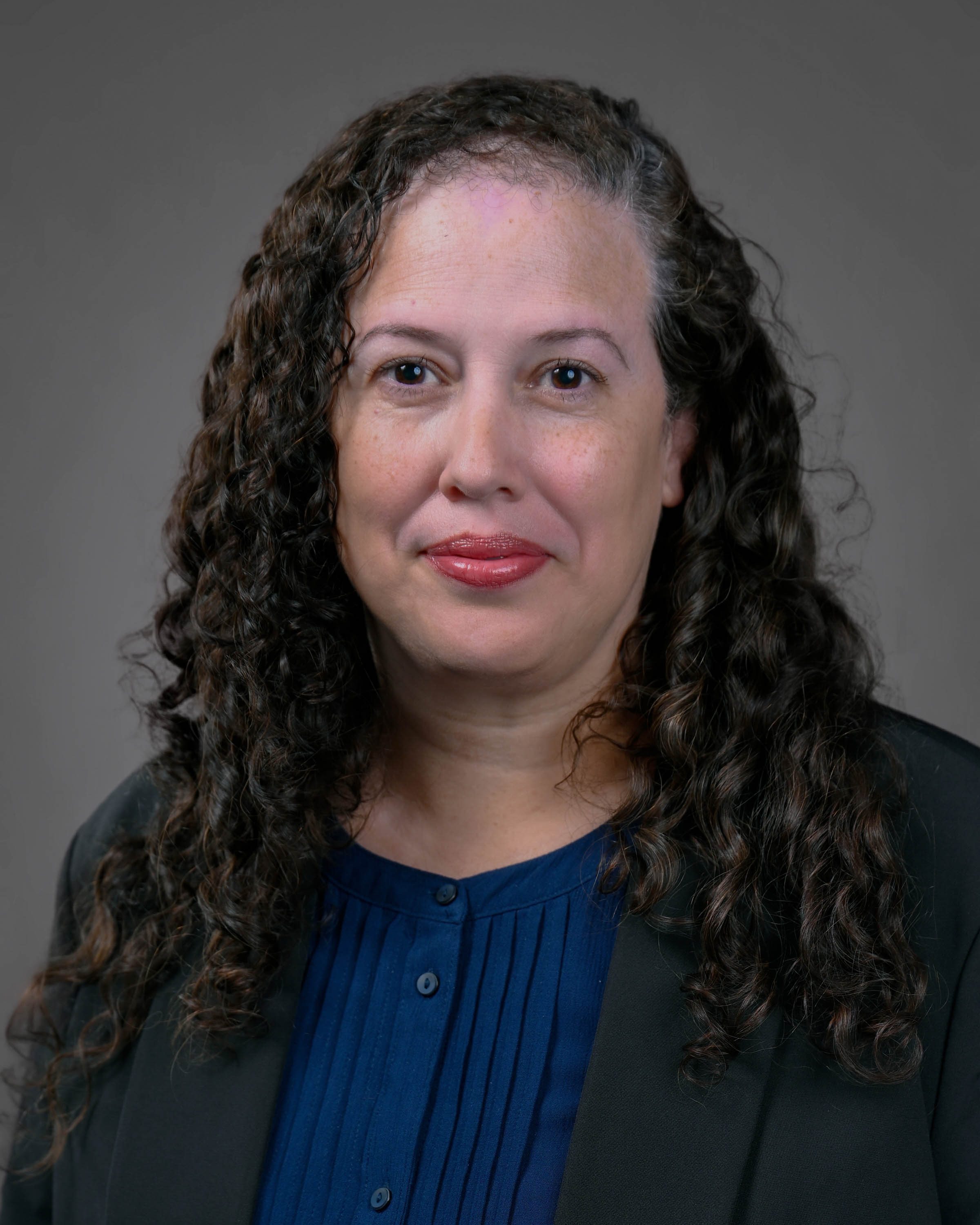 Woman with curly brown hair in a dark suit jacket with a blue shirt.